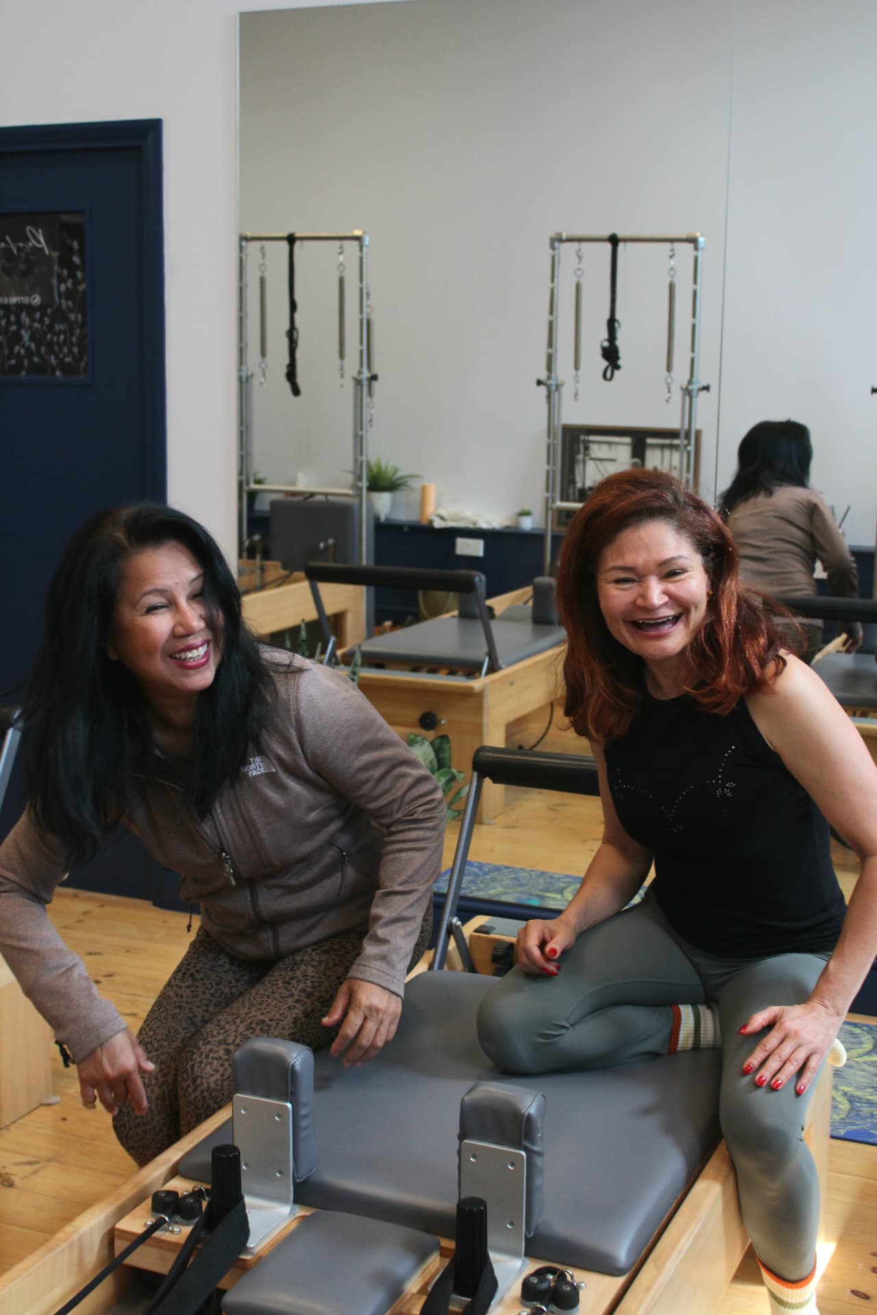 Adriana and Rebecca laughing together while sitting on a Reformer carriage
